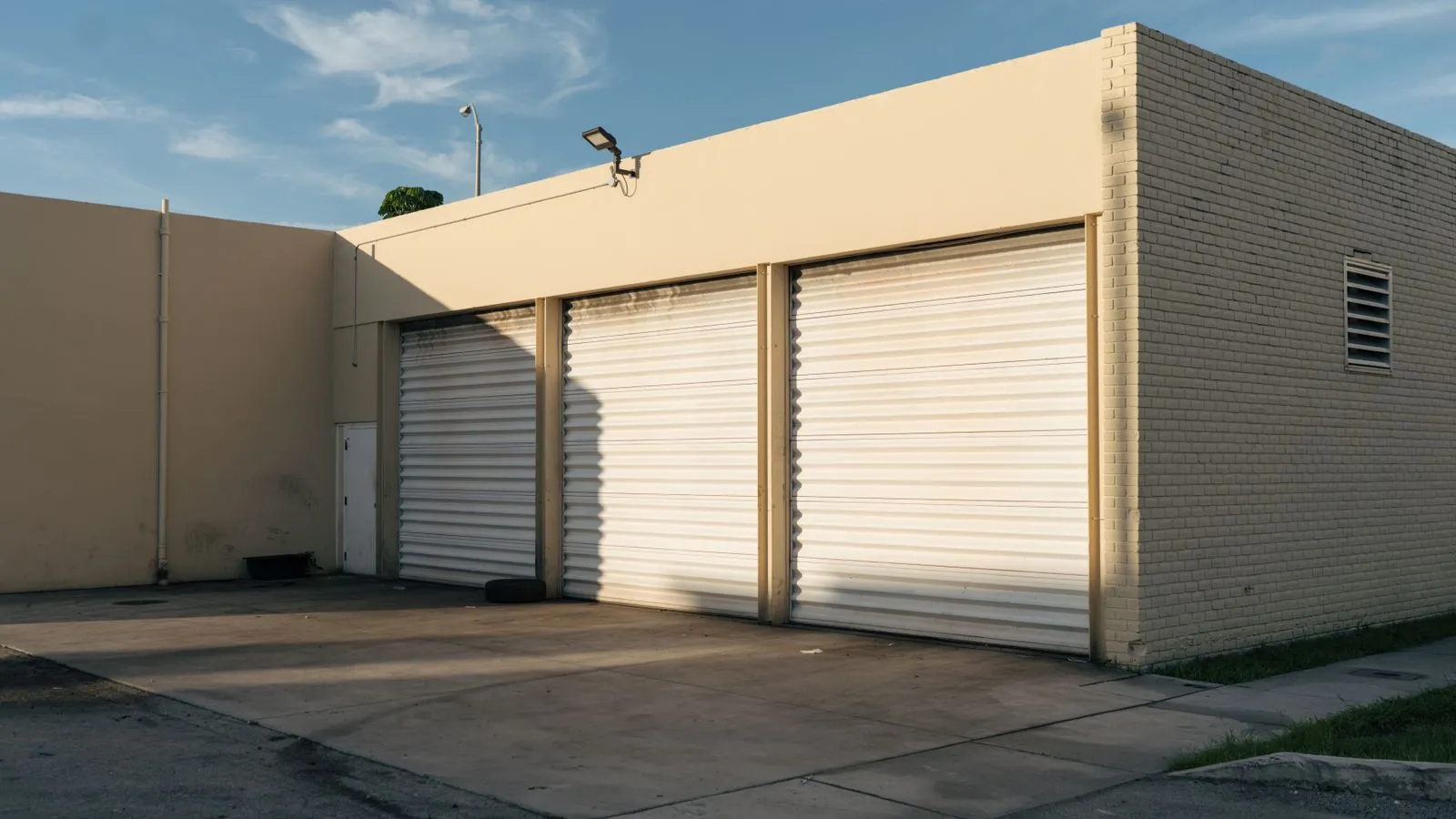 Exterior view of a beige industrial building with three closed garage doors under a clear sky.